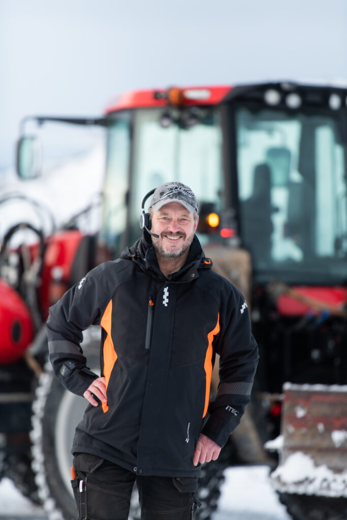 Smiling man with a headset and a hat in front of a red excavator in the snow