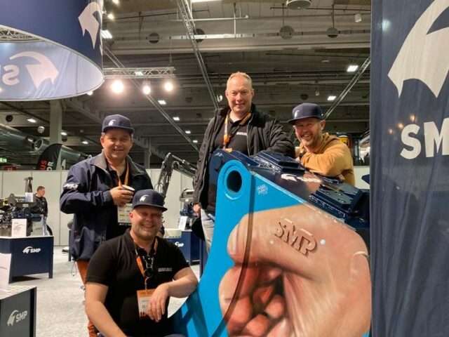 Four happy men in next to a painted excavator bucket from SMP in an exhibition hall