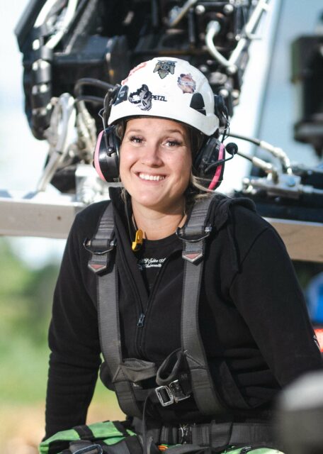 Woman smiling with a hard hat and safety equipment in front of an excavator