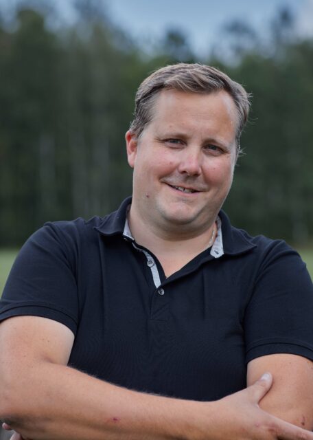 Smiling man with a black shirt outside on a field next to a forest