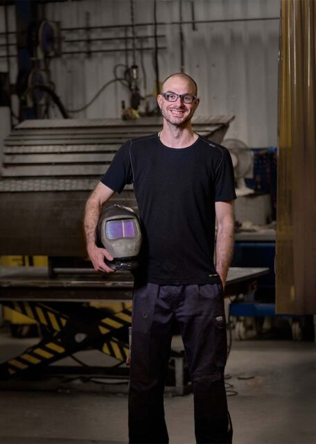 Welder holding his welder hat smiling in front of a large excavator bucket