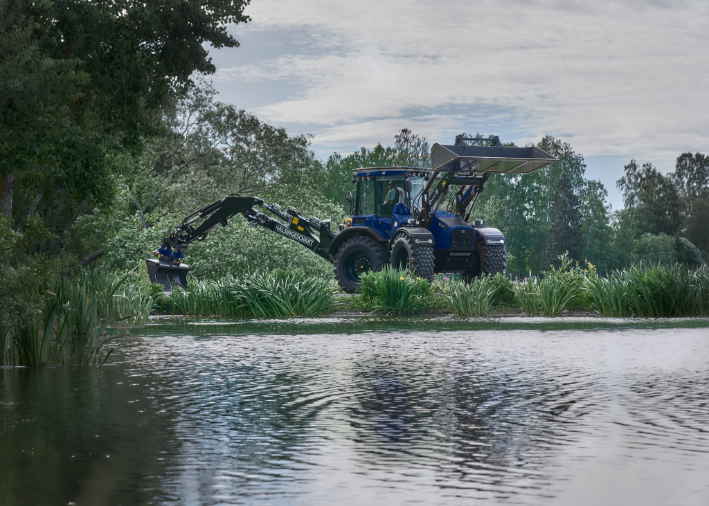 Blue tractor with two buckets attached doing some work in front of a pond